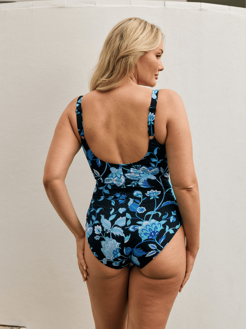Back of model standing by wall wearing a scoop back tank one piece in floral print
