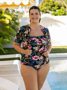 A woman wearing a tropical floral print two-piece swimsuit stands by a swimming pool. The set features a short-sleeve button-up shirt and matching high-waisted briefs with a dark navy background and pink, green, and white floral motifs.