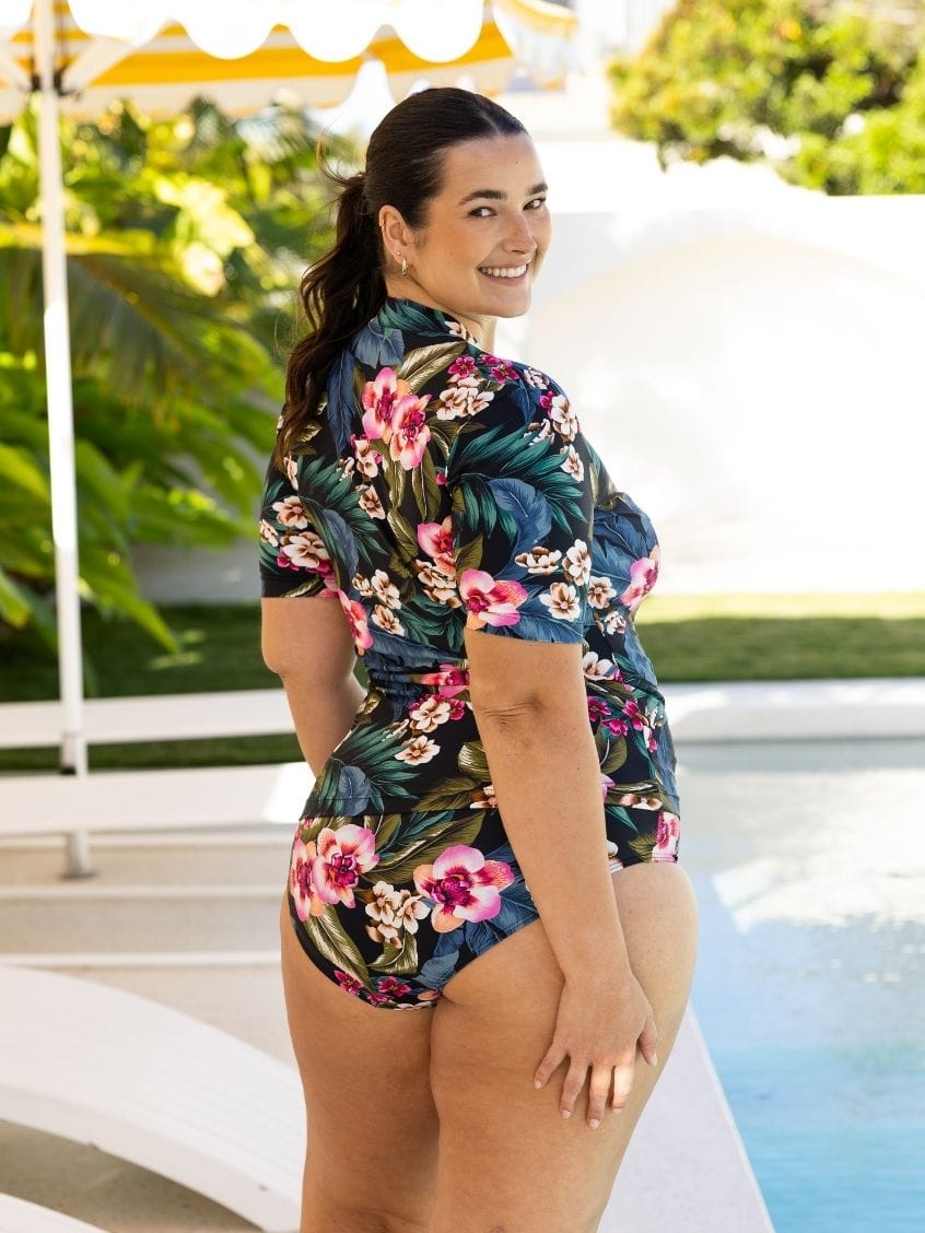 A woman wearing a short-sleeve floral one-piece swimsuit stands by a swimming pool, looking back over her shoulder. The swimsuit has a dark navy base with pink, red, and green tropical floral patterns.