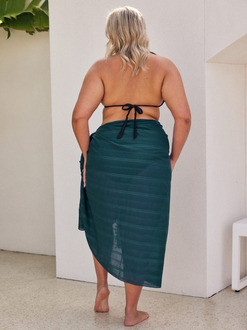 Woman wearing a green sarong standing against a white wall.