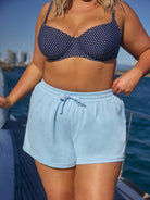 Person wearing a navy polka dot bikini top and light blue shorts on a boat with water and cityscape in the background.