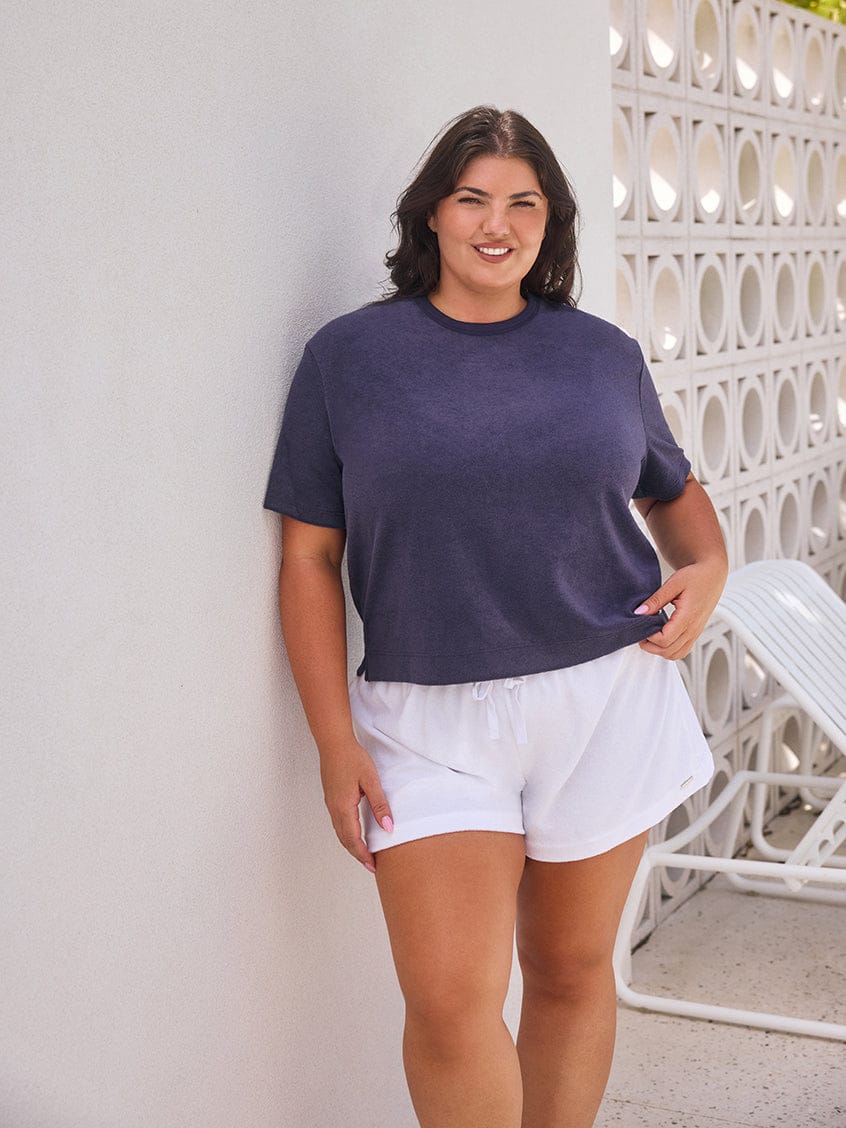 Woman wearing a dark blue t-shirt and white shorts standing against a light-colored wall.