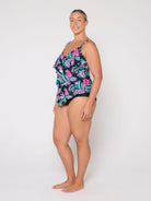 A woman modeling a 3 tier tankini with a tropical leaf pattern in pink, teal, and green on a black background, photographed against a plain white studio backdrop.
