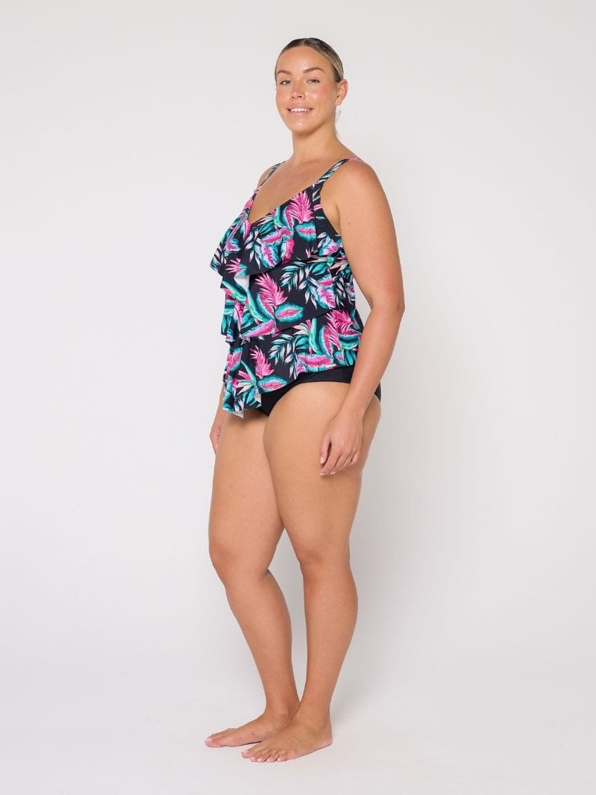 A woman modeling a 3 tier tankini with a tropical leaf pattern in pink, teal, and green on a black background, photographed against a plain white studio backdrop.
