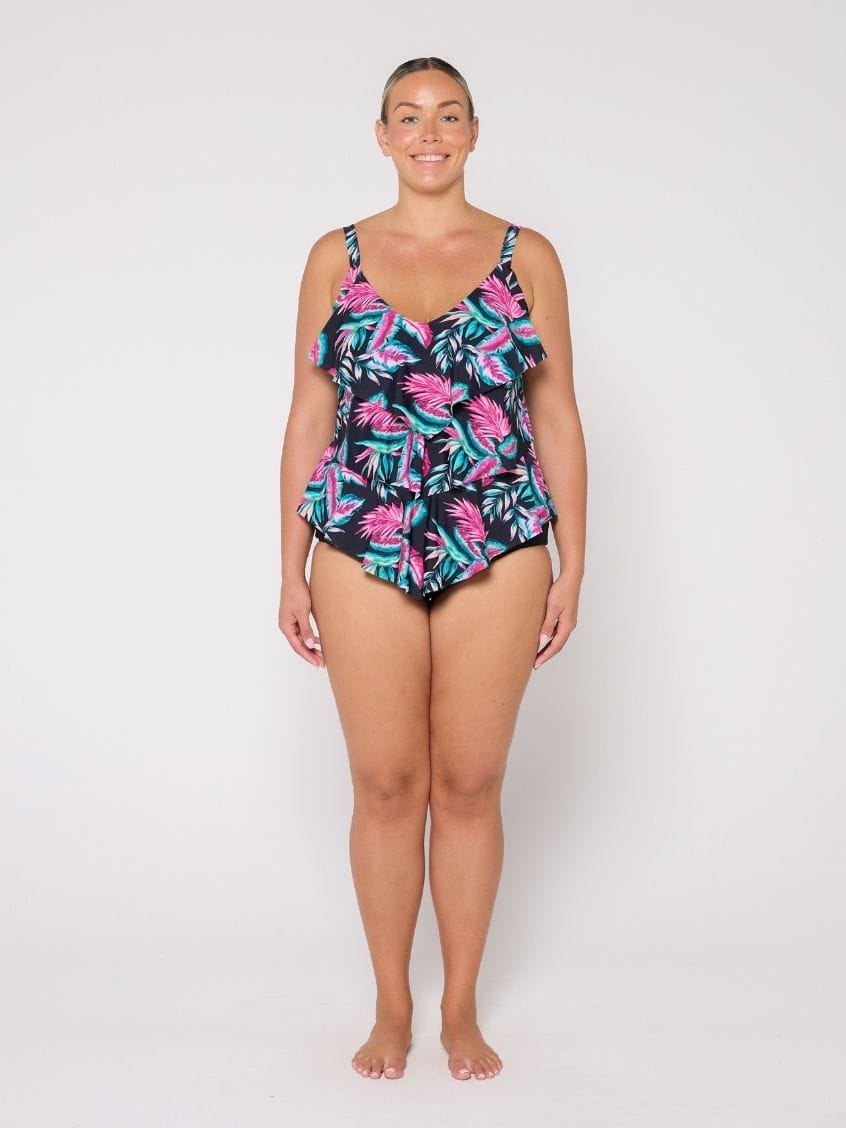 A woman modeling a 3 tier tankini with a tropical palm leaf pattern in pink, teal, and black against a plain white studio background.