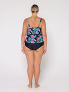 A woman viewed from the back wearing a 3 tier tankini  with a black base and a colorful tropical leaf pattern in pink, teal, and green, paired with solid black bottoms, against a plain white studio background.
