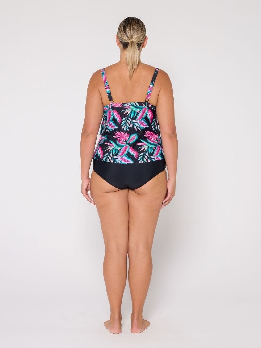 A woman viewed from the back wearing a 3 tier tankini  with a black base and a colorful tropical leaf pattern in pink, teal, and green, paired with solid black bottoms, against a plain white studio background.