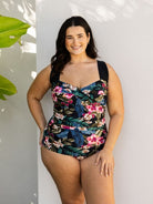 A woman modeling a one-piece swimsuit with a dark tropical floral print, standing against a light gray wall with leaf shadows.