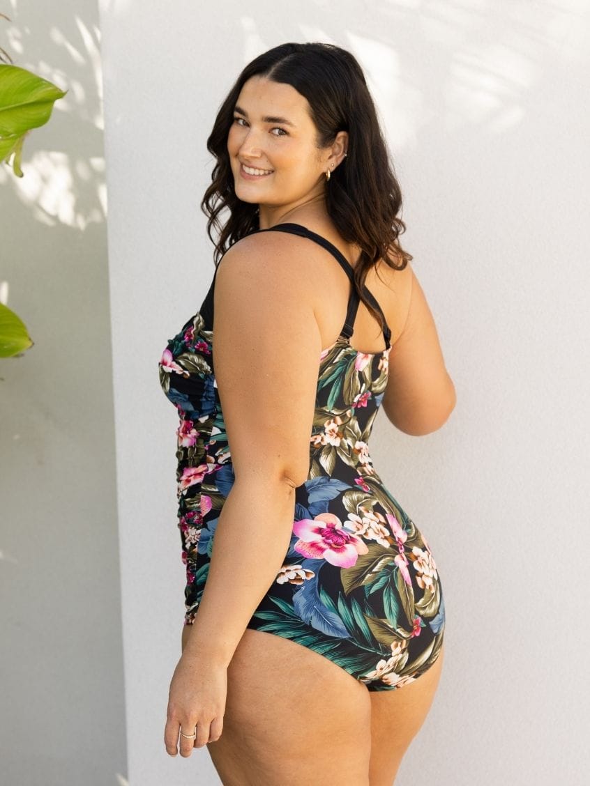 A woman modeling a one-piece swimsuit with a tropical floral print, photographed against a white wall with soft leaf shadows.