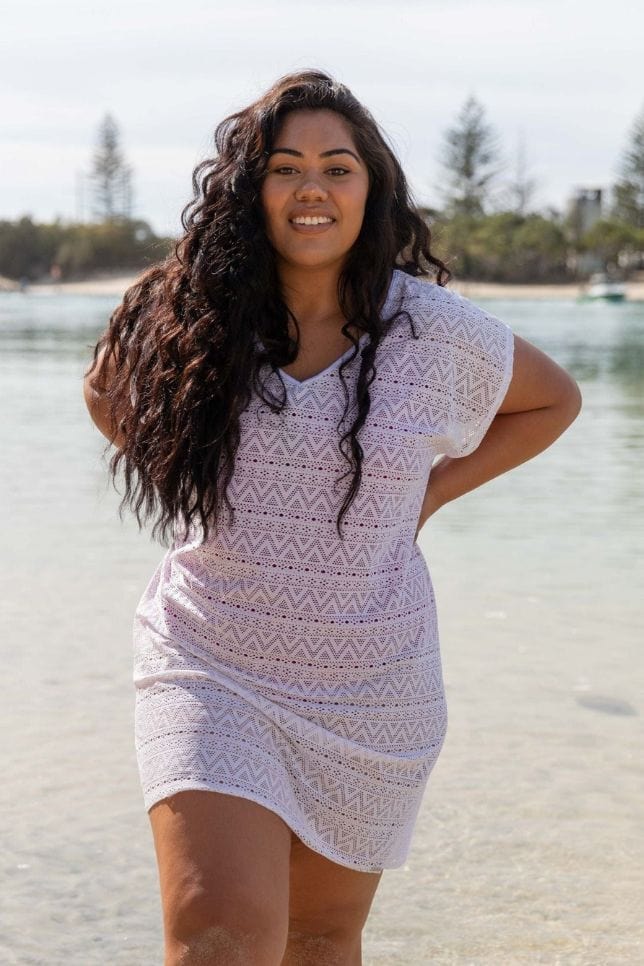 Woman stands on beach in a white kaftan. The beach cover up is made of a white mesh and has small patterned cut-outs in the mesh