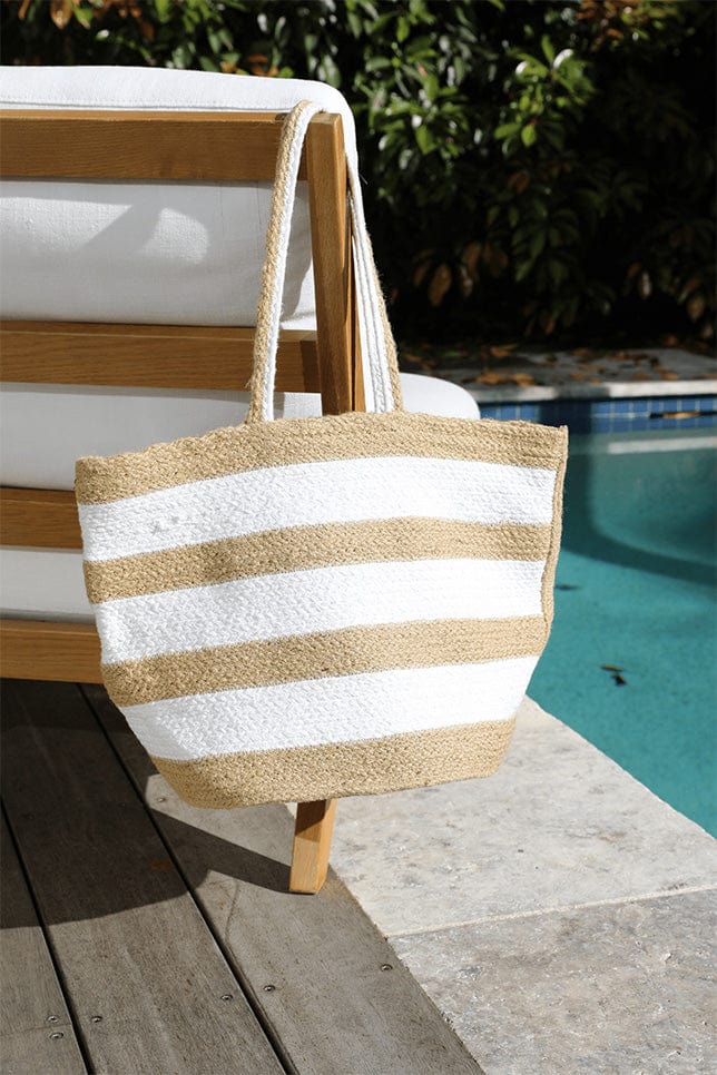 beach bag hangs on a chair by the pool. The bag has alternating white and beige/ natural horizontal stripes