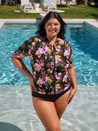 Brunette model wears stunning pink and gold floral short sleeve rash vest in chlorine resistant fabric with front zip