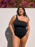 Woman wearing a black one-shoulder swimsuit standing against a light background with purple flowers.