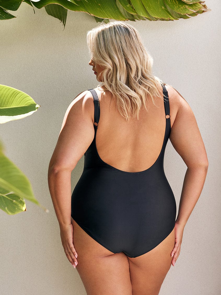 back of model standing in front of a white wall wearing a black one piece swimsuit