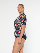 Blonde model shows the side of pool proof rash vest with short sleeve in a black based floral print