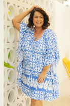 brunette model leaning against a white wall wearing a cornflower blue and white block print beach dress