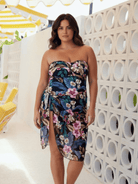 A woman models a strapless floral wrap dress with a sweetheart neckline and a thigh-high slit. The dress features a vibrant tropical floral pattern in shades of navy, teal, pink, and green.