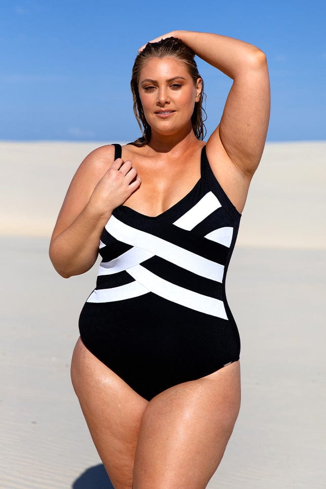 Plus size model stands on sand dunes in a black and white one piece swimsuit with criss cross pattern along the torso