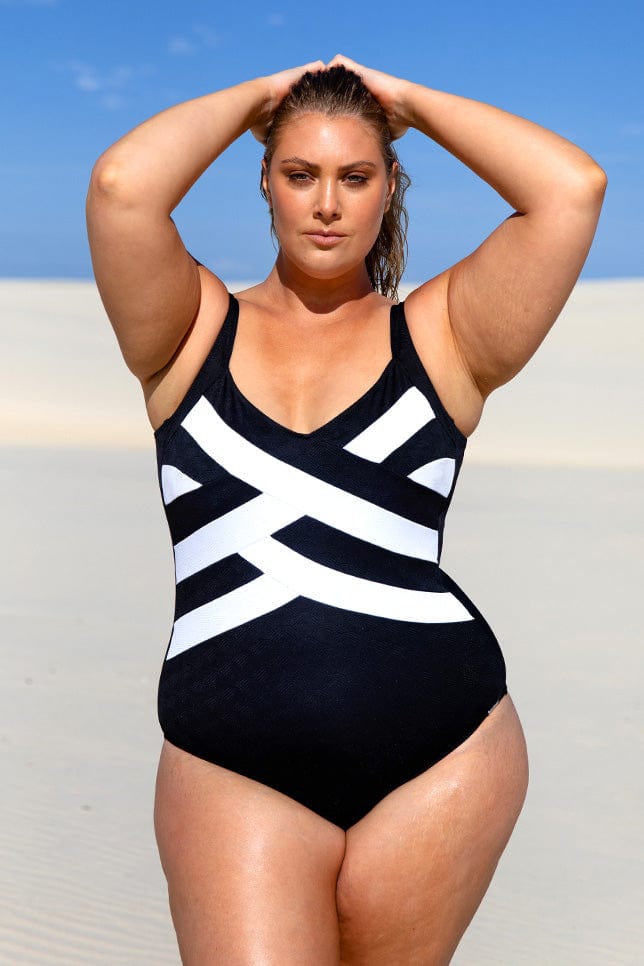 Woman wears a black and white one piece swimsuit with a striped criss cross pattern at the front v-neck.  The model is in her 20s, blonde, she stares into the camera, standing on white sand dunes with blue sky.
