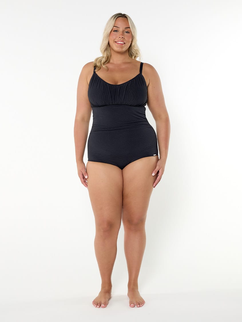 A woman modeling a black one-piece swimsuit with thin shoulder straps and a gathered sweetheart neckline, standing against a plain white studio background.