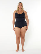 A woman modeling a black one-piece swimsuit with thin shoulder straps and a gathered sweetheart neckline, standing against a plain white studio background.