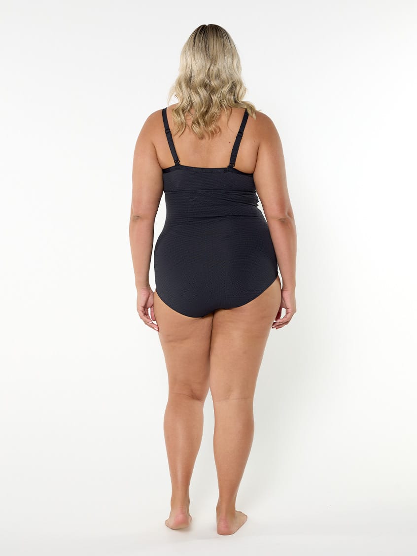 A woman viewed from the back wearing a black one-piece swimsuit, standing against a plain white studio background.