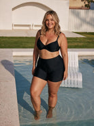 A woman modeling a black two-piece swimwear set consisting of a bandeau-style bikini top and high-waisted shorts, standing in shallow water next to a pool in an outdoor backyard setting.