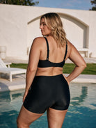 A woman stands by a swimming pool wearing a black two-piece swimwear set consisting of a backless strappy bikini top and high-waisted briefs.