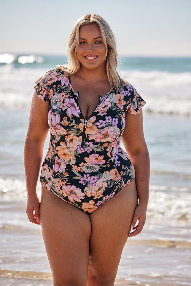 Blonde model wearing black floral zip front swimsuit at beach