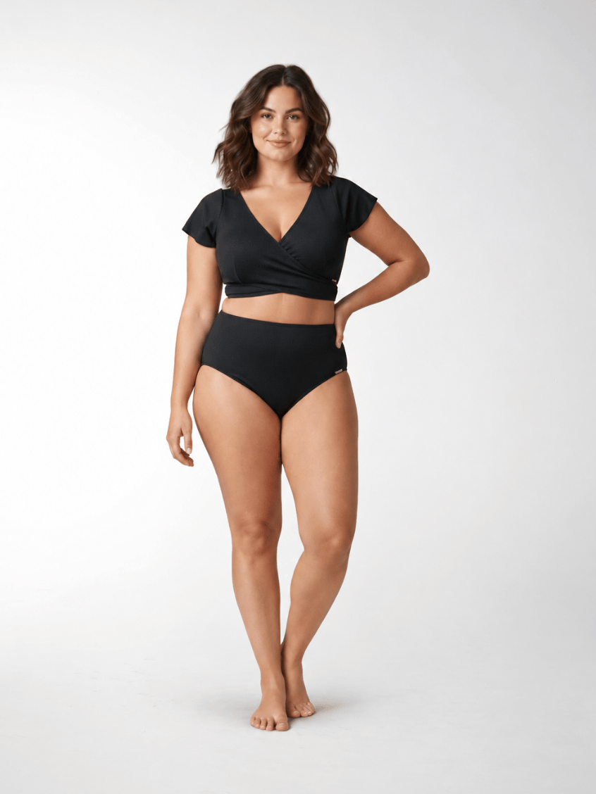 A woman modeling a black two-piece swimsuit consisting of a short-sleeve wrap-front crop top and matching high-waisted briefs, photographed against a seamless light gray studio background.