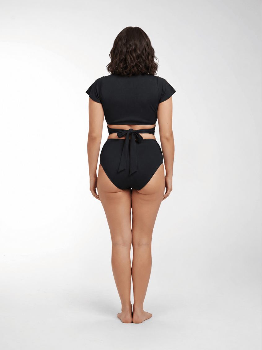 A woman stands with her back to the camera wearing a black two-piece swimsuit with a short-sleeve crop top and high-waisted bottoms featuring a tie detail at the waist, against a light gray studio background.