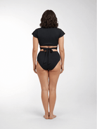 A woman stands with her back to the camera wearing a black two-piece swimsuit with a short-sleeve crop top and high-waisted bottoms featuring a tie detail at the waist, against a light gray studio background.