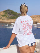 Woman wearing a white t-shirt with 'La Dolce Vita' design by a waterfront with boats and green hills.
