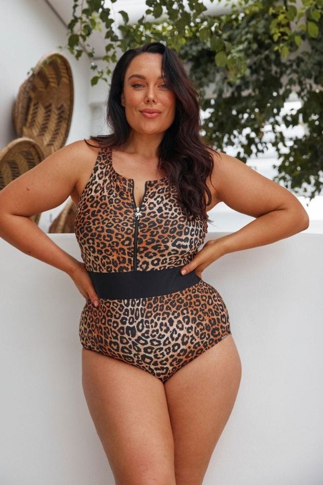 Dark haired woman wears a leopard print swimsuit with a black waist band. The one piece is sleeveless with a zipper along the front.