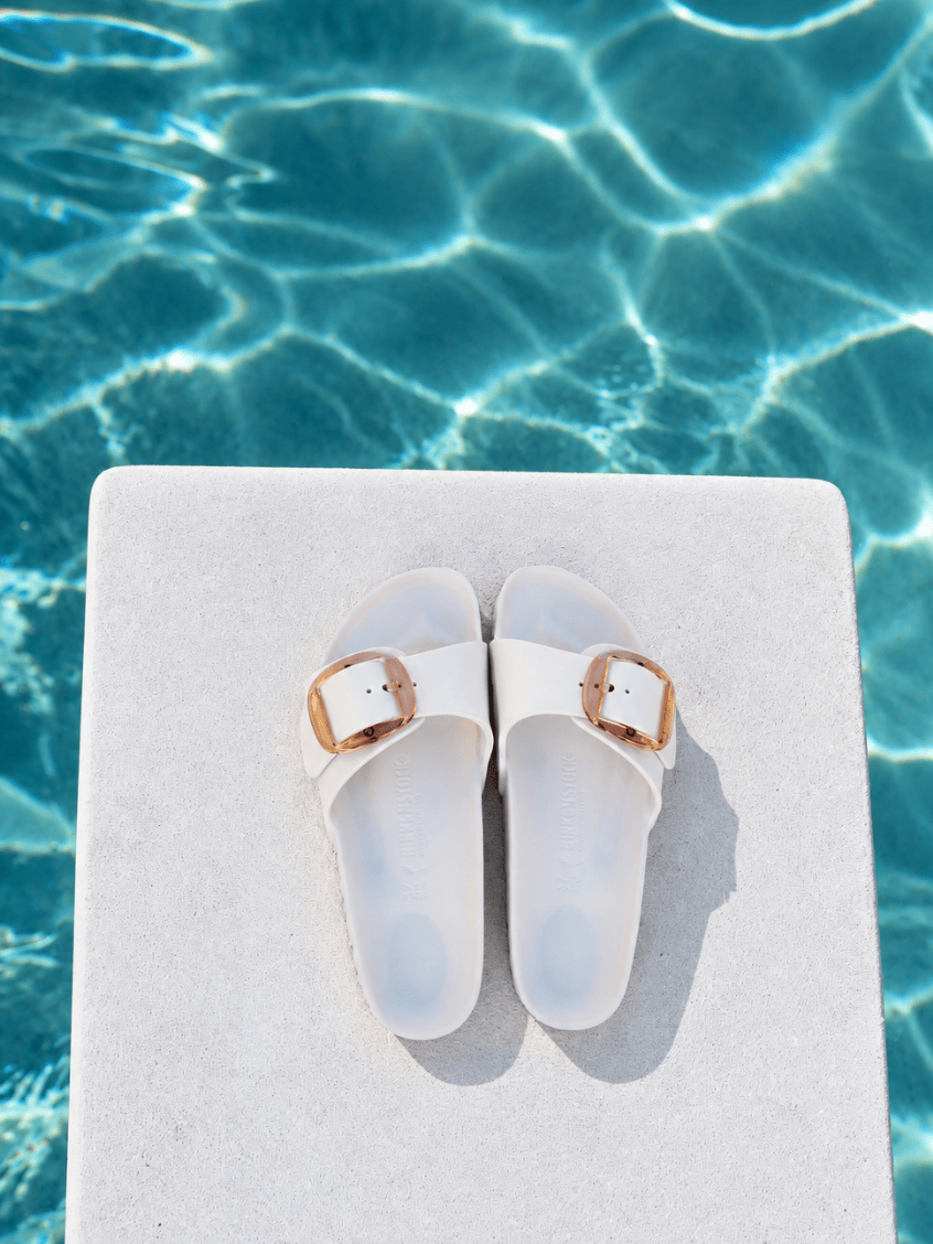 A pair of white slide sandals with rose gold buckle details, resting on a white stone slab with a swimming pool background.