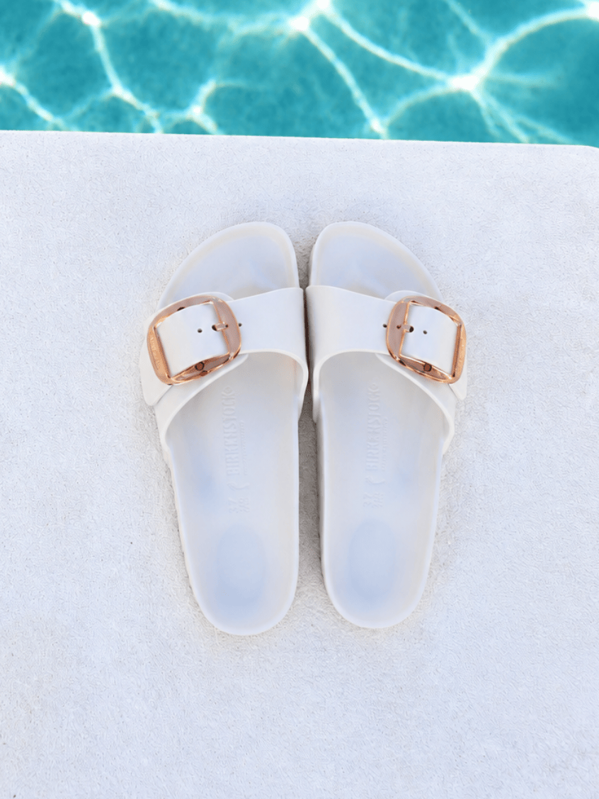A pair of white slide sandals with metallic rose gold buckle straps, laid flat on a white textured surface with a swimming pool background visible at the top.