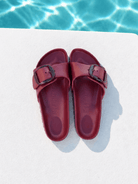 A pair of burgundy double-strap slide sandals with dark metal buckles, laid flat on a white deck with a swimming pool background visible at the top.