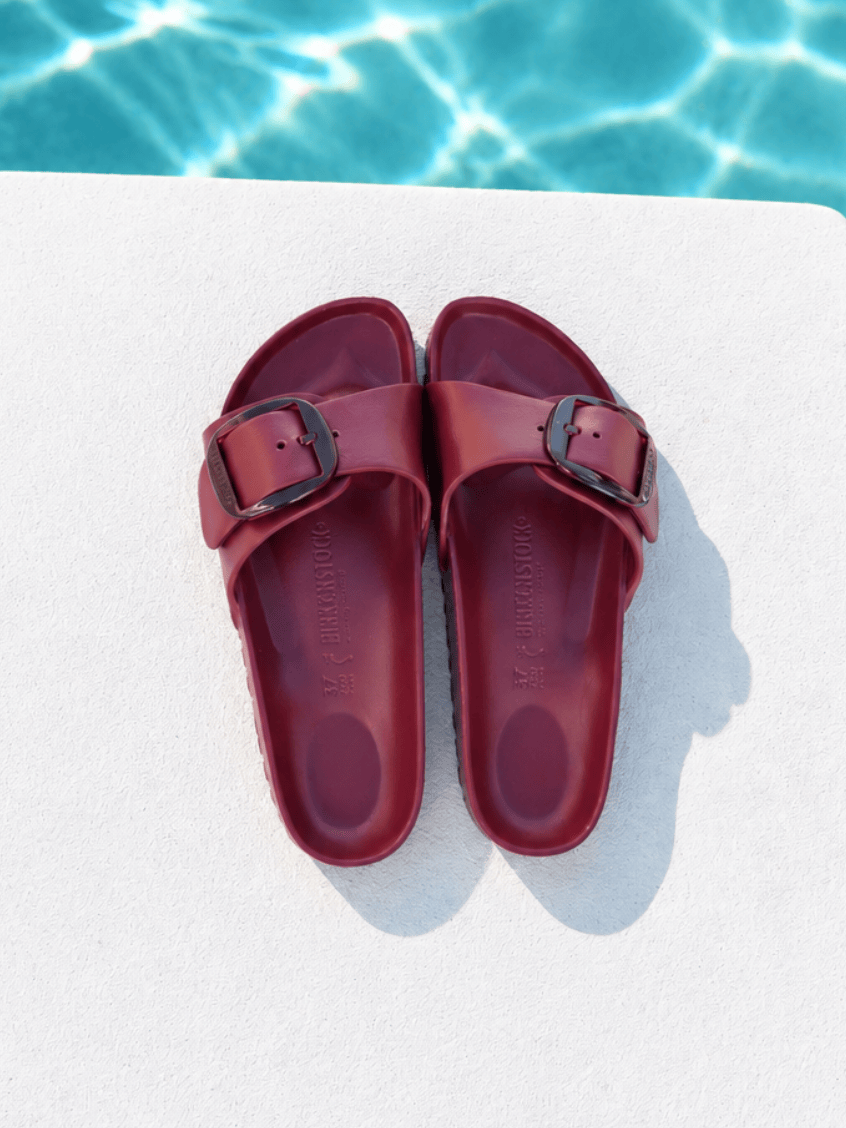 A pair of burgundy double-strap slide sandals with dark metal buckles, laid flat on a white deck with a swimming pool background visible at the top.