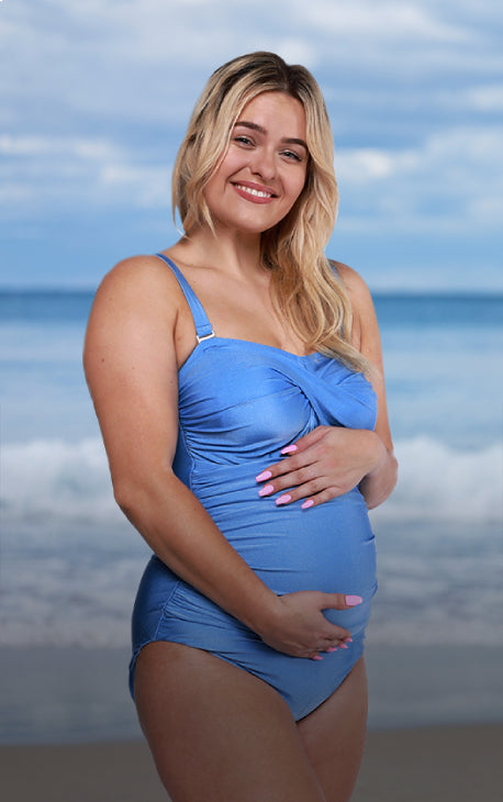 Pregnant model cradles her stomach, wearing Capriosca Swimwear's maternity range blue bandeau one piece.