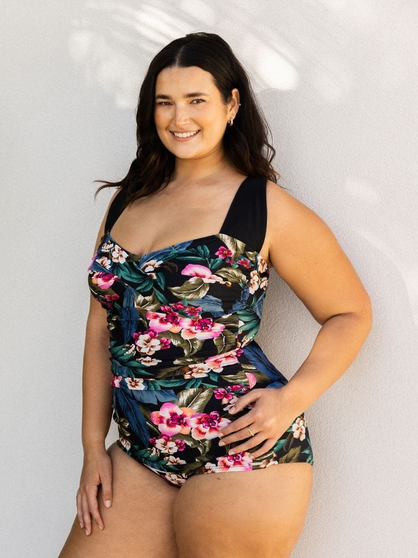 A woman modeling a one-piece swimsuit with a dark navy and black base featuring a vibrant pink, red, and green tropical floral print, posed against a light gray textured wall.