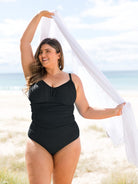 A woman stands on a sandy beach wearing a black underwire one-piece swimsuit and holding a white scarf overhead.