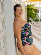 A woman wearing a strapless one-piece swimsuit with a tropical floral print leans against a white wall in an outdoor poolside setting with lounge chairs and green plants in the background.