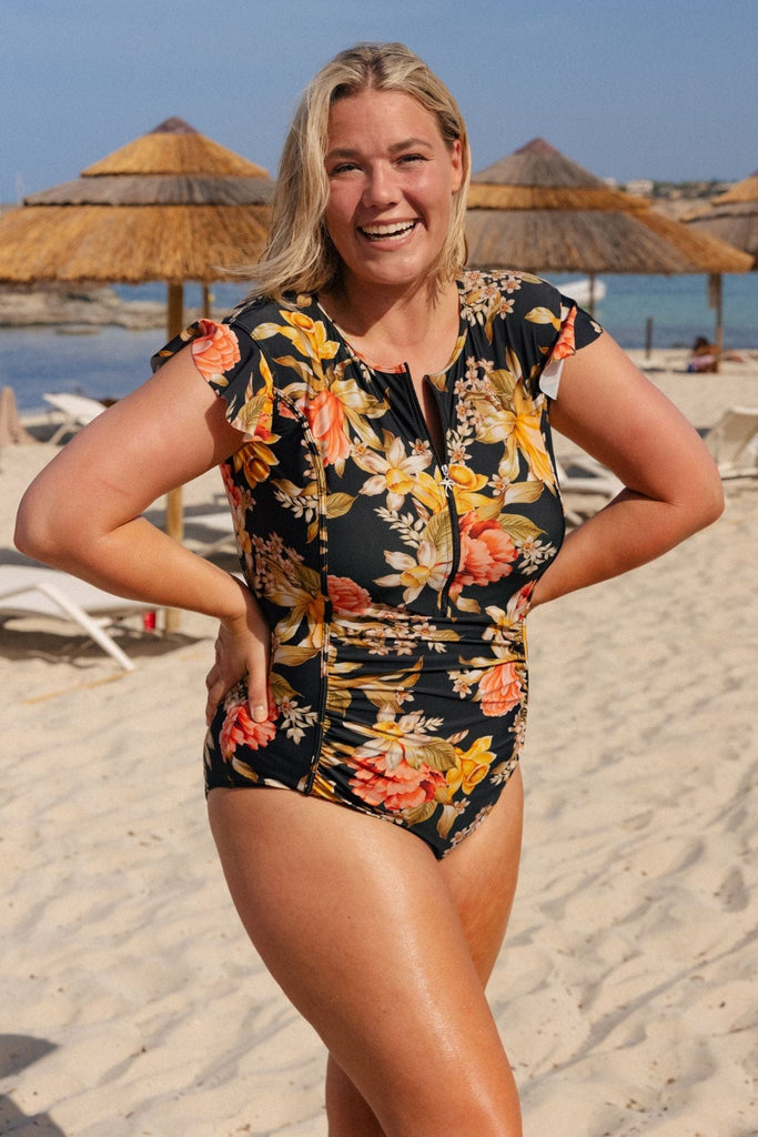 Blonde model on beach wearing a one piece swimsuit with frill sleeve in black, yellow, orange print